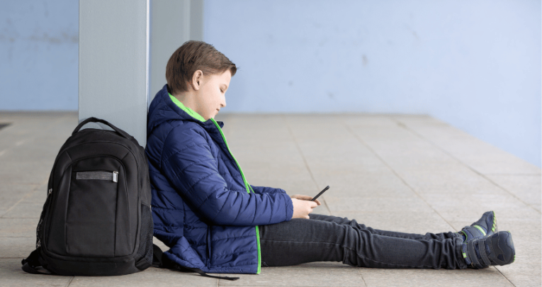 Boy with schoolbag sitting on floor looking at phone, representing poor school attendance