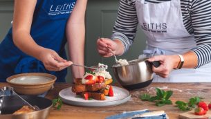 A teacher helps a child in a Leiths Academy apron to prepare a dessert using strawberries and cream