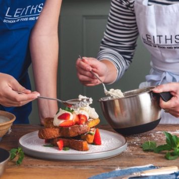 A teacher helps a child in a Leiths Academy apron to prepare a dessert using strawberries and cream