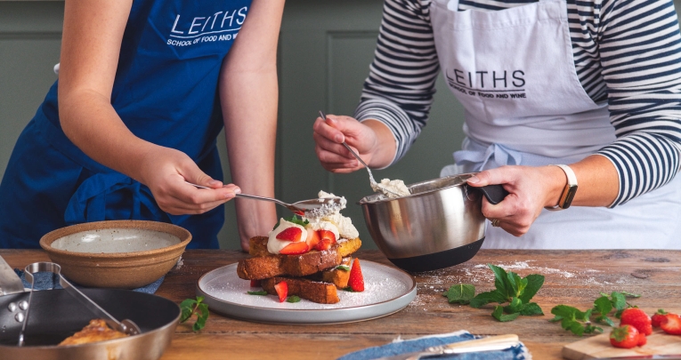 A teacher helps a child in a Leiths Academy apron to prepare a dessert using strawberries and cream