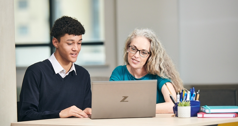 A male secondary school student sitting at a laptop with a tutor