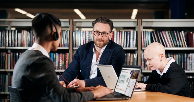 Two secondary school students sitting at a desk in a library with a teacher